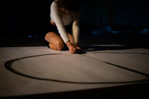 A woman drawing a large circle on the floor in a dimly lit space, focusing on artistic expression.