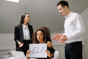A woman in an office holds a 'HELP' sign while facing conflict with coworkers.